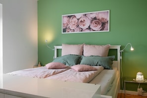 Ein Bett mit weißem Kopfteil, Bettwäsche in Grün und Rosa, und ein an der Wand hängendes Bild von Blumen.