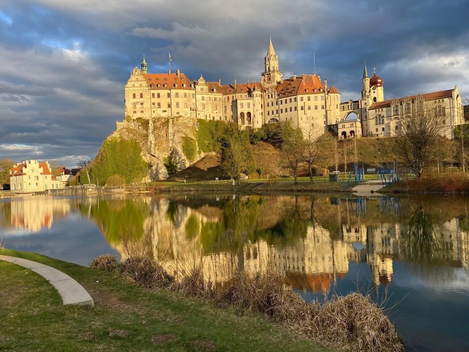 Ein historisches Gebäude mit Spiegelung in ruhigem Wasser, umgeben von Grün und einem klaren Himmel.