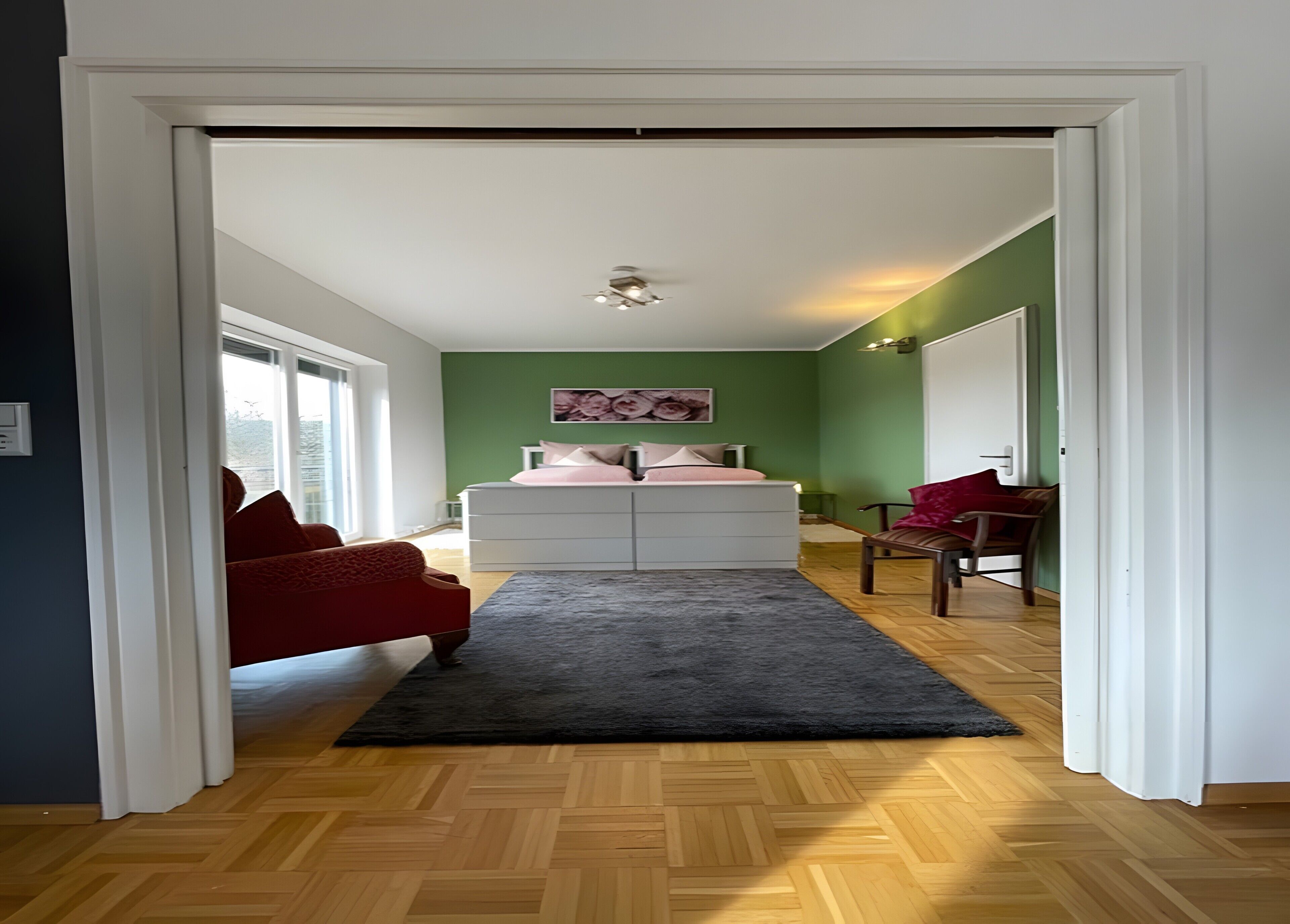 Un couloir mène à une chambre qui contient un lit, une commode, un fauteuil rouge, un banc et une fenêtre.