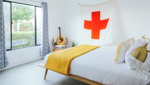 Une chambre avec un lit, une guitare, une décoration murale représentant une croix rouge et une vue sur la verdure à l’extérieur.