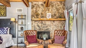 Una habitación con pared de piedra, chimenea, dos sillones de cuero, cama, estante con objetos decorativos y ventana con cortinas.