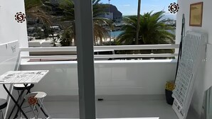 A balcony with a table and chairs, a white radiator, and a view of a coastal area with buildings and palm trees. - Sea view studio with balcony, 2 steps from the beach (Mogán)