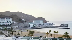 A coastal cityscape with a beach, buildings, and a hilly background. - Sea view studio with balcony, 2 steps from the beach (Mogán)