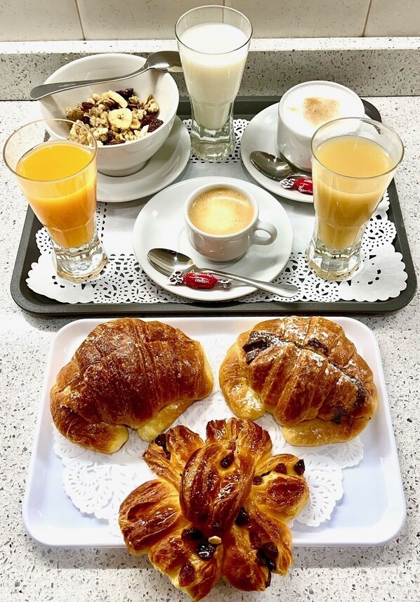 Una bandeja de desayuno con cruasanes, un tazón de cereal con frutas, un vaso de jugo de naranja, un vaso de leche, una taza de café y un vaso de jugo de naranja.