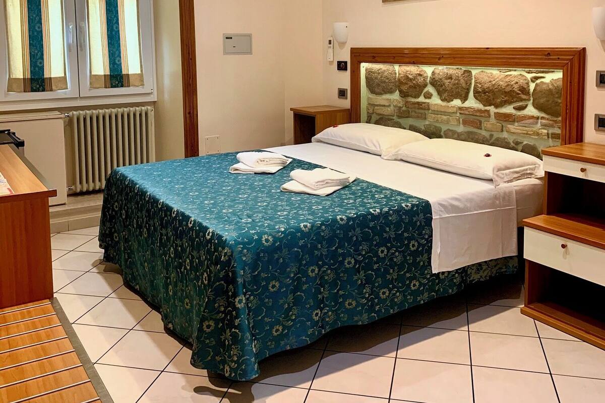 Une chambre d’hôtel comprenant un lit, une table de chevet, une commode en bois, un radiateur, une fenêtre avec des rideaux et un tableau accroché au mur.