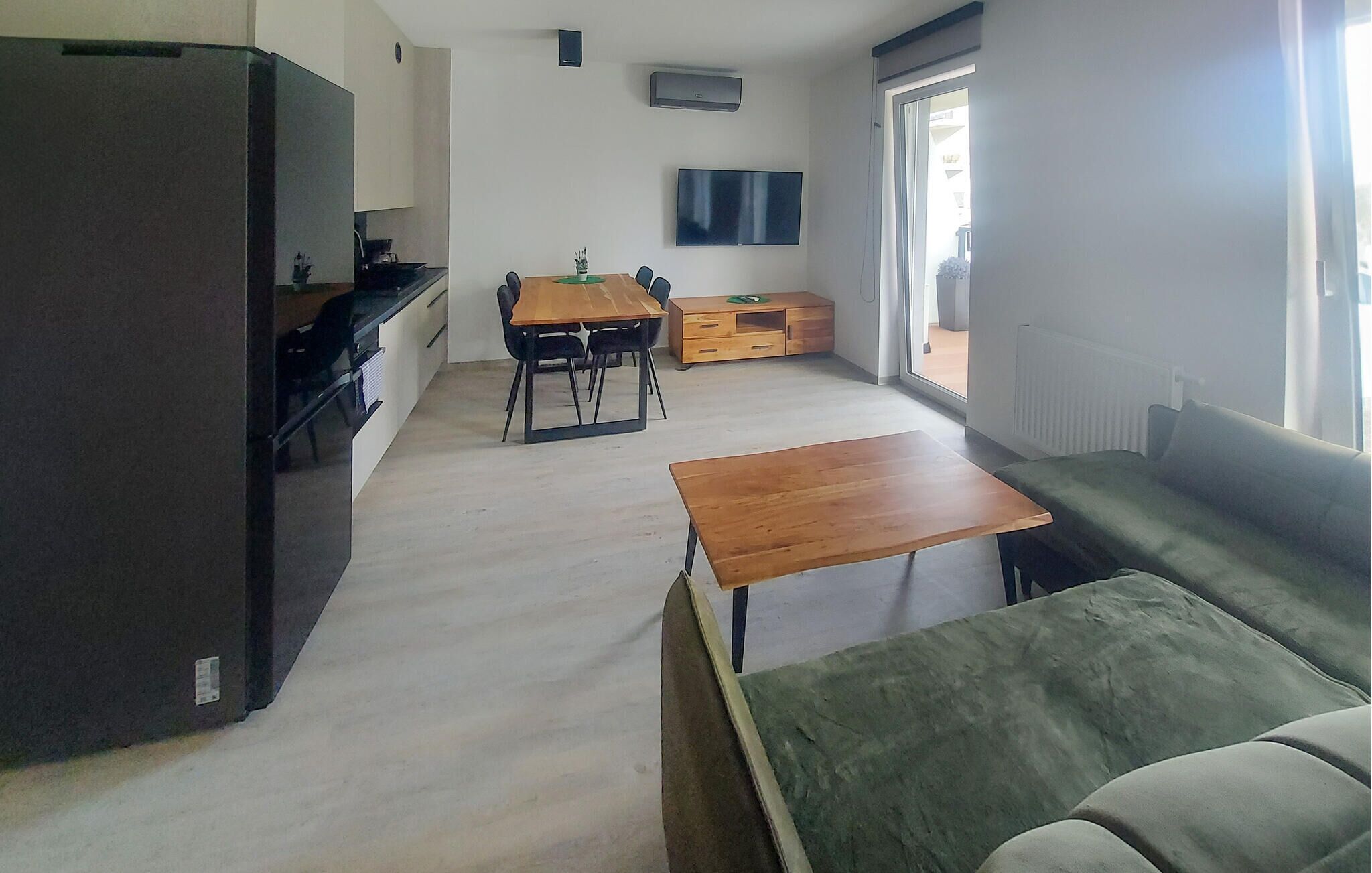 Una sala de estar moderna con un sofá, una mesa de centro de madera, una mesa de comedor con sillas, una zona de cocina con un refrigerador y un televisor montado en la pared.