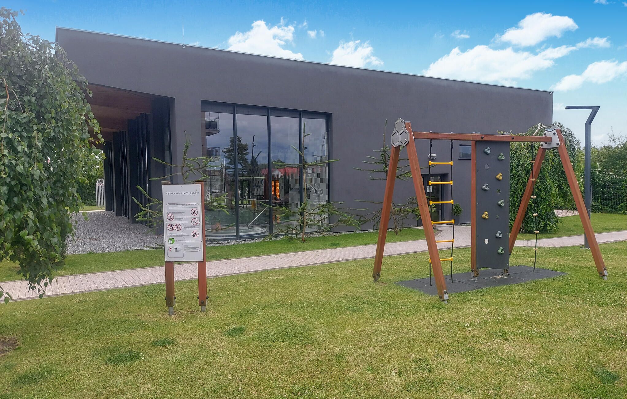 Un edificio moderno con entrada de vidrio, una estructura de escalada de madera y un cartel frontal.