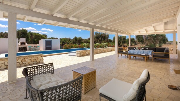 An outdoor patio with wicker furniture, a stone table, and a view of a swimming pool and greenery. - 4134 Villa Majer by Perle di Puglia (Manduria)