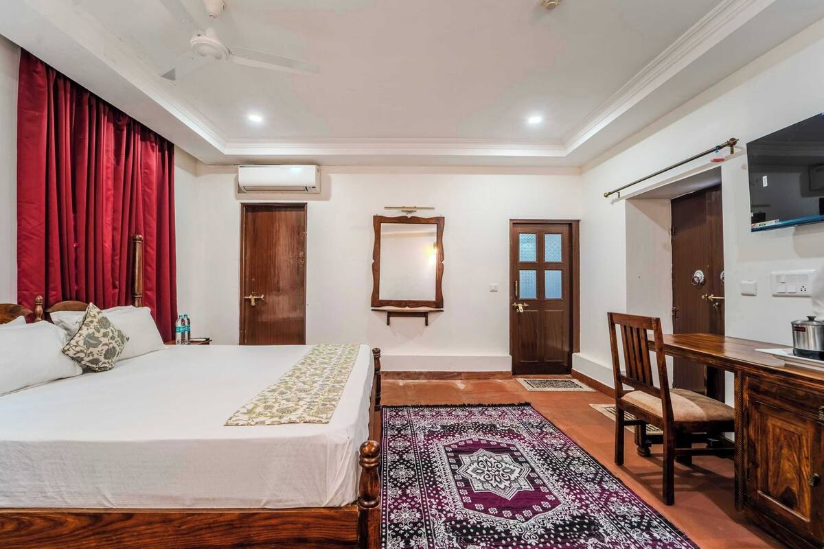 A hotel room with a bed, a wooden desk, a chair, a mirror, and a red curtain.