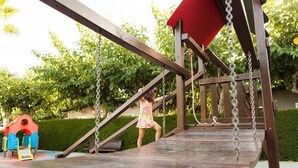 A wooden swing set with a red canopy and a small playhouse in the background.