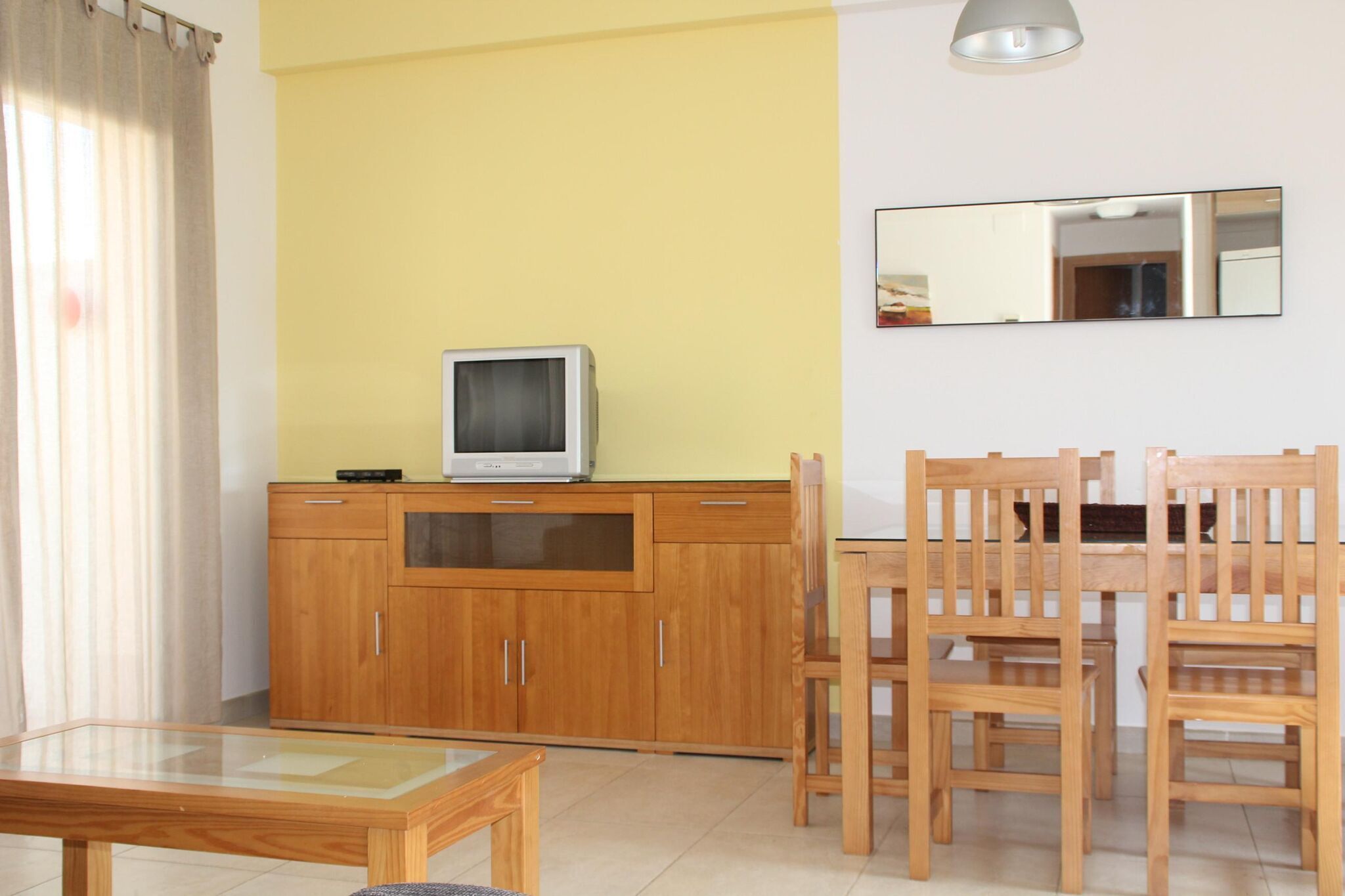 A room with a wooden cabinet, a television on top, a dining table with chairs, and a mirror on the wall.