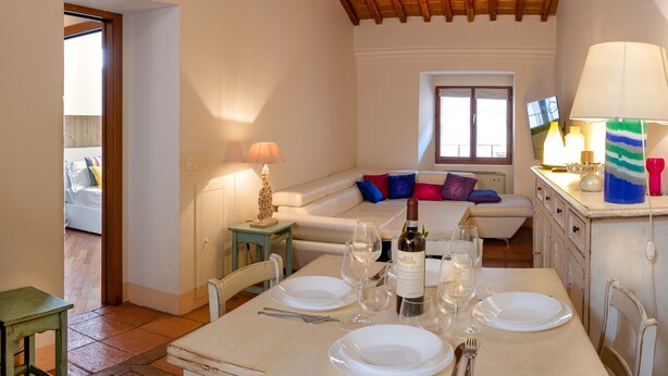 A dining area with a table set for two, a sofa with colorful cushions, and a television on the wall. - Il Castellare, a delightful apartment in the heart of Sierna (Siena)