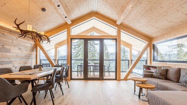 A spacious room with a high ceiling, wooden walls, a large dining table with chairs, a sofa, and a view through a glass door to the outdoors. - Appealing chalet with finnish sauna (Turrach)