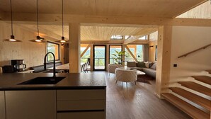 A modern kitchen with a central island, wooden flooring, and pendant lighting. A dining area with a sofa and chairs is visible through a glass door. - Awesome chalet with sauna (Turrach)