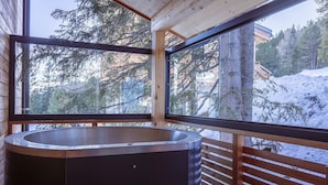 Um deck de madeira com uma banheira de hidromassagem grande, guarda-corpos de madeira e vista para árvores cobertas de neve e uma cabana.