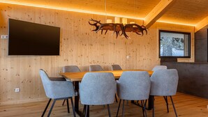 Uma sala de reuniões moderna com mesa de madeira e cadeiras cinzas, uma TV na parede e teto de madeira com um lustre de chifres exclusivo.