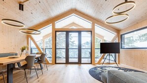 A modern interior with a wooden ceiling, a dining area with chairs and a table, a sofa, and a large window with a view of trees.