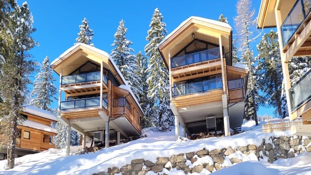 A modern ski resort building with multiple balconies and wooden exterior, surrounded by snow-covered trees.