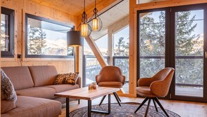 Ein gemütliches Zimmer mit Holzbalkendecke, großen Fenstern, einer Couch, einem Tisch und zwei Stühlen.