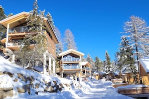 Un albergue de montaña cubierto de nieve con varios niveles y grandes ventanales, rodeado de árboles cargados de nieve.