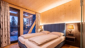 A bedroom with a double bed, wooden walls, a glass door leading to a balcony, and a nightstand with a lamp. - Lavish chalet with sauna (Turrach)