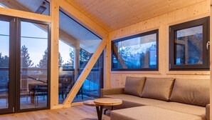 A cozy cabin interior with a wooden floor, a sofa, and a table. Large windows offer views of the outdoors. - Lavish chalet with sauna (Turrach)