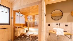 A wooden sauna with benches and a glass door, a sink with a mirror, and a window with blinds. - Lavish chalet with sauna & hot tub (Turrach)