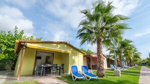 A yellow building with a covered patio, two blue lounge chairs, and a palm tree. - Bungalow in Sant Salvador near Beaches (Tarragona)