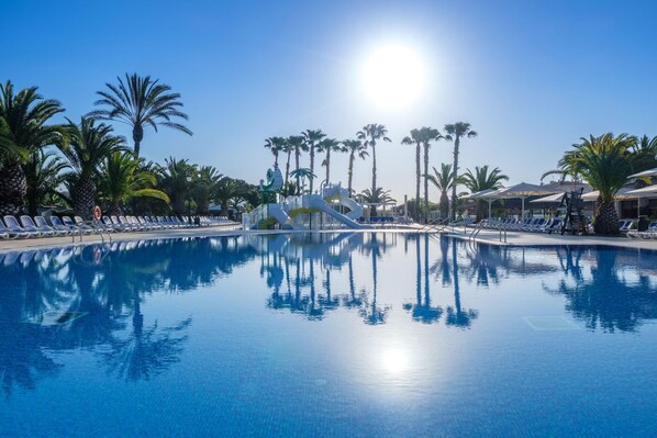 Una piscina azul cristalina con un tobogán, rodeada de palmeras y sillas de descanso.