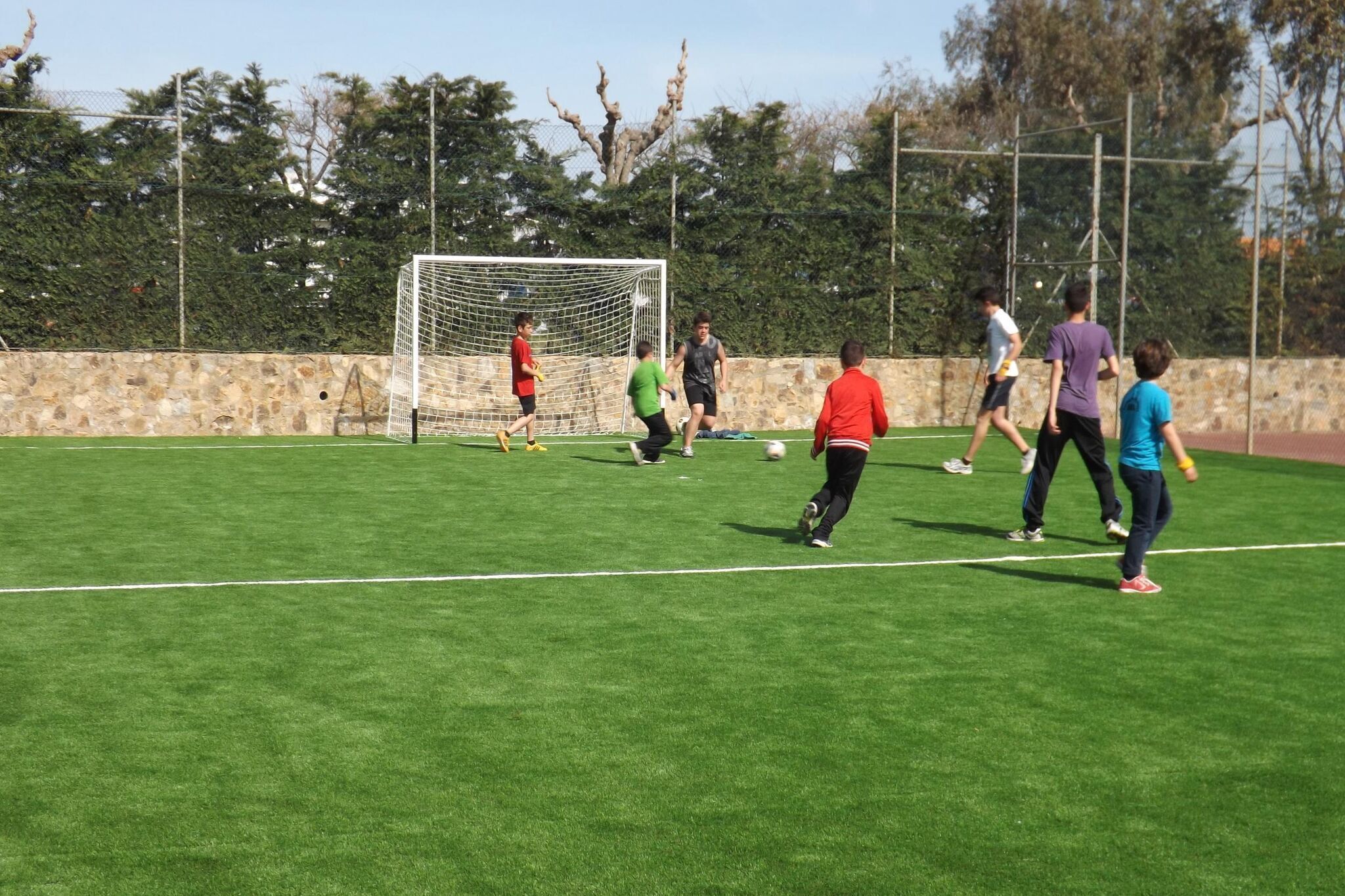 A soccer field with a goalpost, players, and a stone wall in the background.