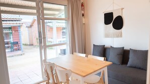 A room with a wooden table and chairs, a grey sofa with cushions, a large window with curtains, and a wall-mounted fire extinguisher. - Bungalow in Malgrat near Golden Beaches (Barcelona)