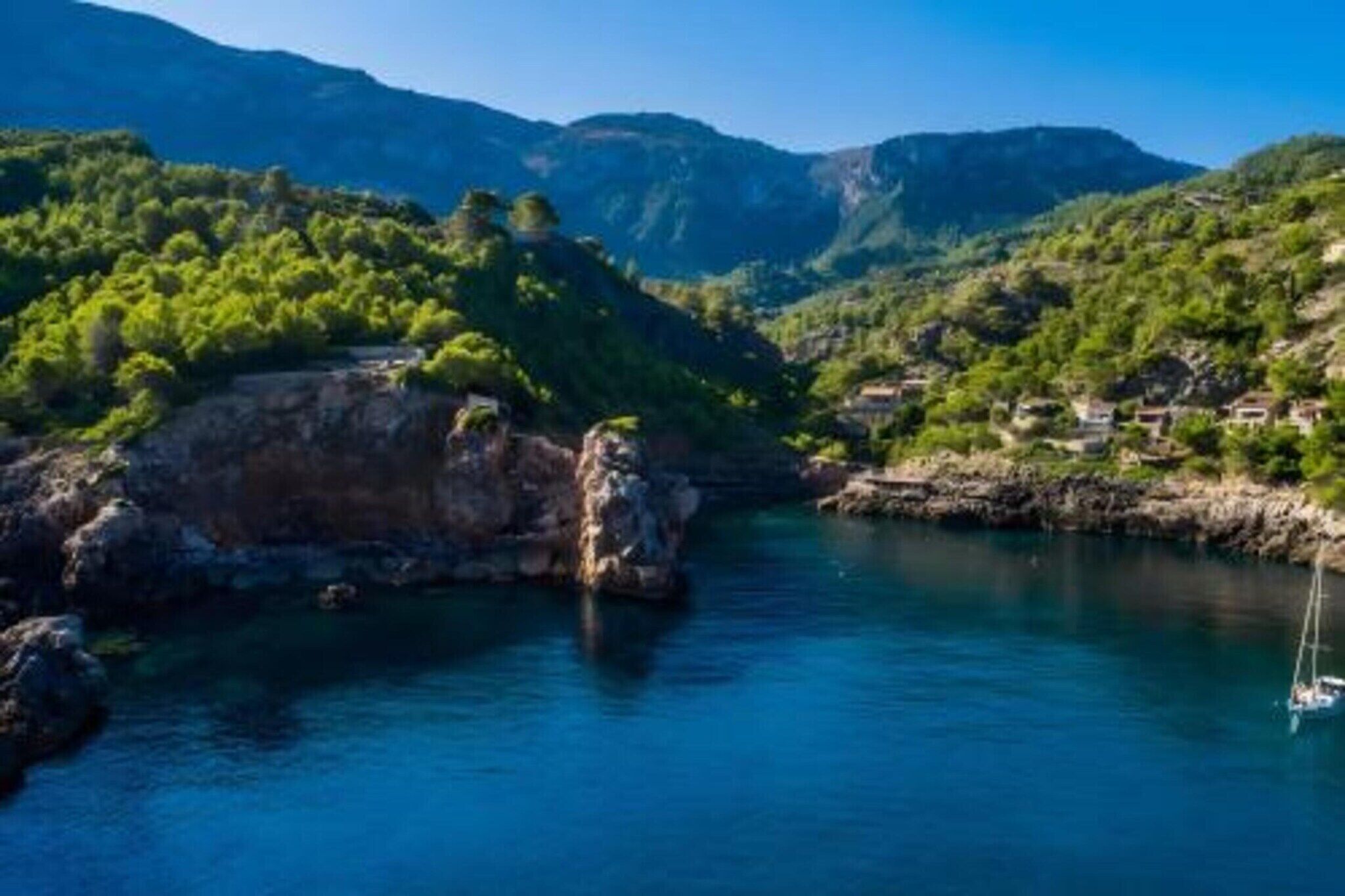 A coastal landscape with a rocky outcrop, clear blue water, and a sailboat.