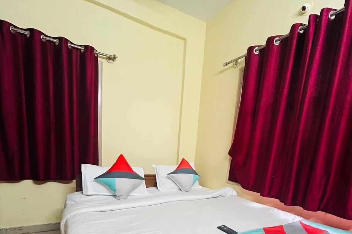 A single bed with a patterned bedspread, two pillows, and red curtains.