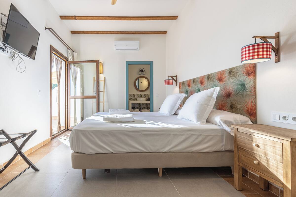 A neatly made bed with white linens, a wooden bedside table with drawers, a wall-mounted TV, and a window with curtains.