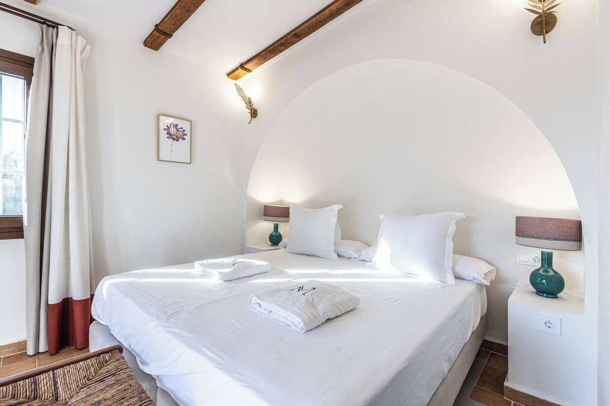 A neatly made bed with white linens and towels, a bedside lamp, and a wall-mounted light fixture.