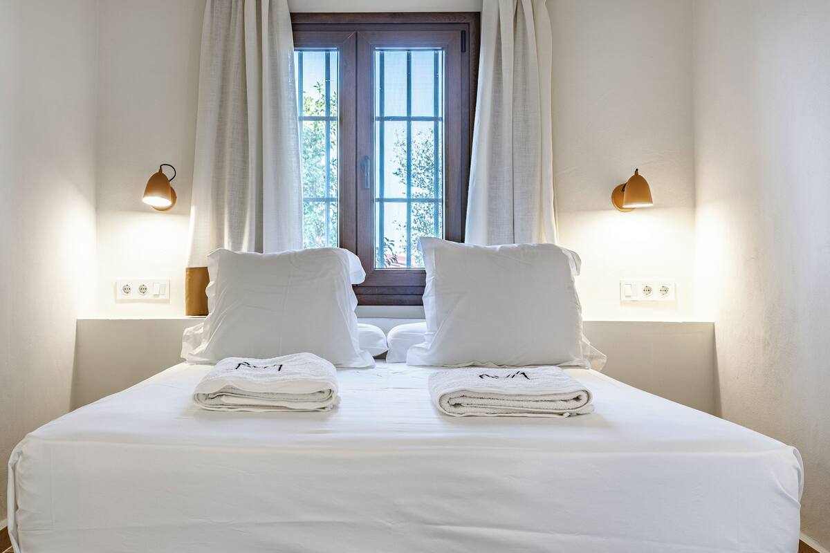 A neatly made bed with white linens and towels, two wall-mounted lamps, and a window with curtains.