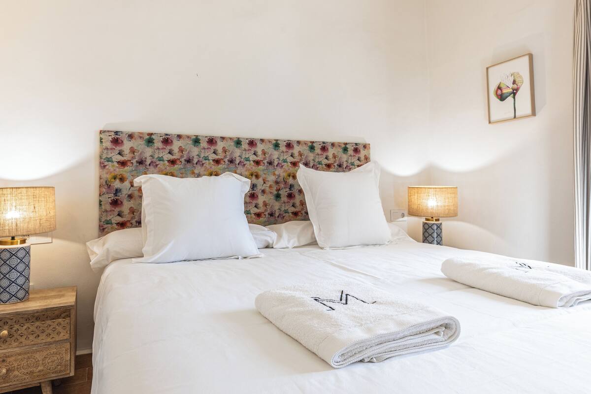 A neatly made bed with white linens and two pillows, a floral headboard, and two bedside lamps on wooden nightstands.