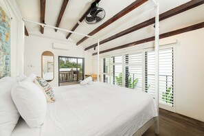 Une chambre avec un grand lit, des poutres en bois, un ventilateur de plafond et une vue sur la verdure par la fenêtre.