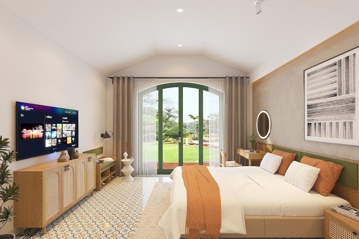 Ein Hotelzimmer mit einem großen Bett, einem an der Wand montierten Fernseher, Blick auf den Garten durch eine Glastür und einem Nachttisch aus Holz mit einer Lampe.