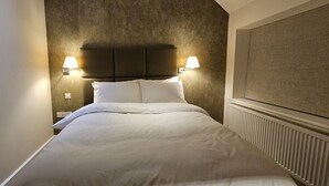 Un lit bien fait, avec du linge de lit blanc, dans une pièce dont le mur présente une texture particulière et qui est agrémenté d’une tête de lit avec deux lampes fixées au mur.