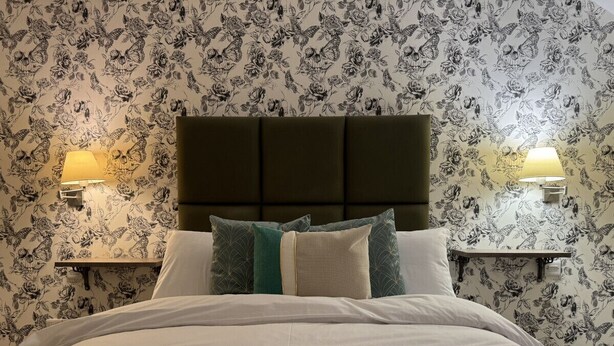 A neatly made bed with a dark headboard, two bedside lamps, and a floral wallpaper pattern. - Class Stays - Room 2 (Shrewsbury)