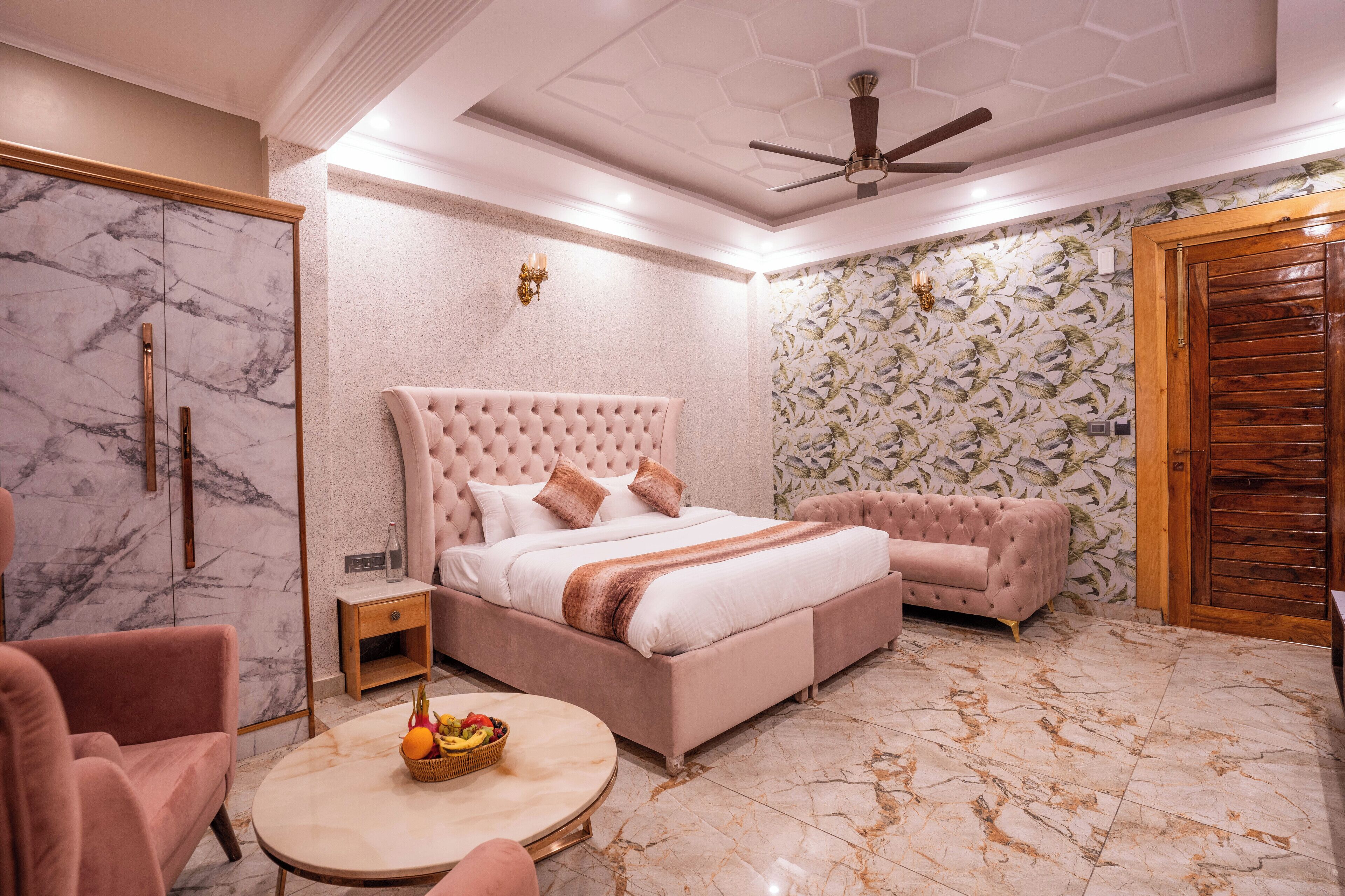 A bedroom with a marble floor, a bed with a tufted headboard, a pink sofa, a round table with fruit, and a wooden door.