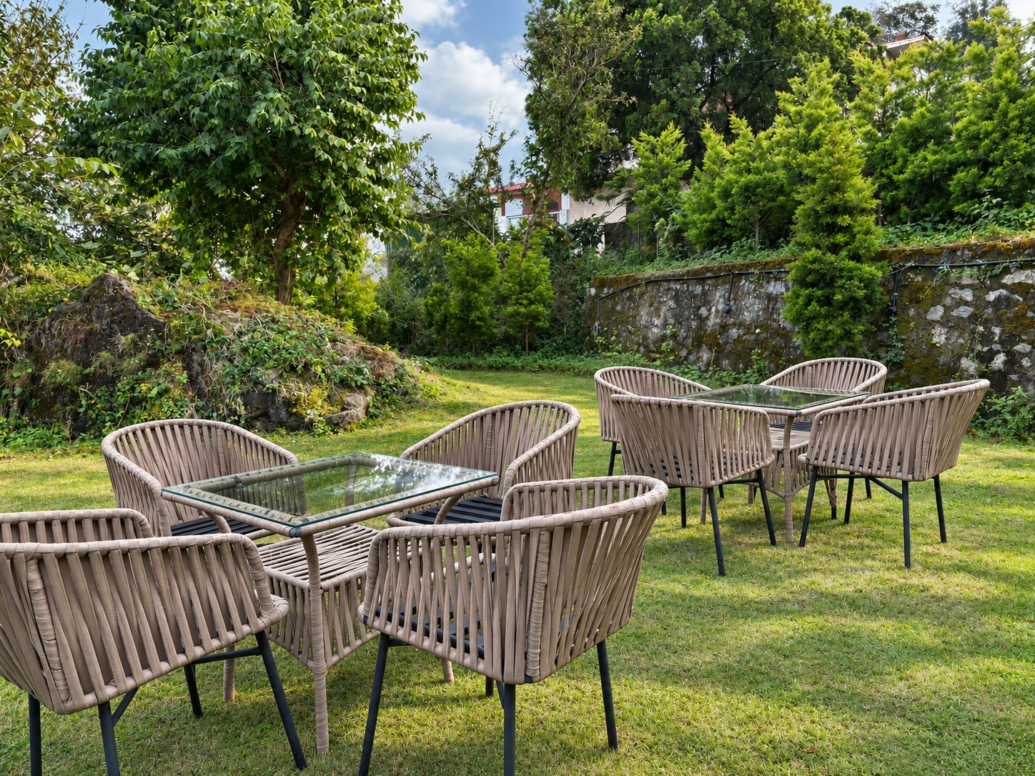 An outdoor seating area with wicker chairs and a glass table, surrounded by lush greenery and a stone wall.