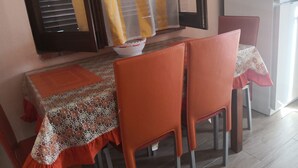 Ruokailutila, jossa on kuviollinen pöytäliina peittämässä pöytää, oranssit tuolit ja taustalla valkoinen jääkaappi.