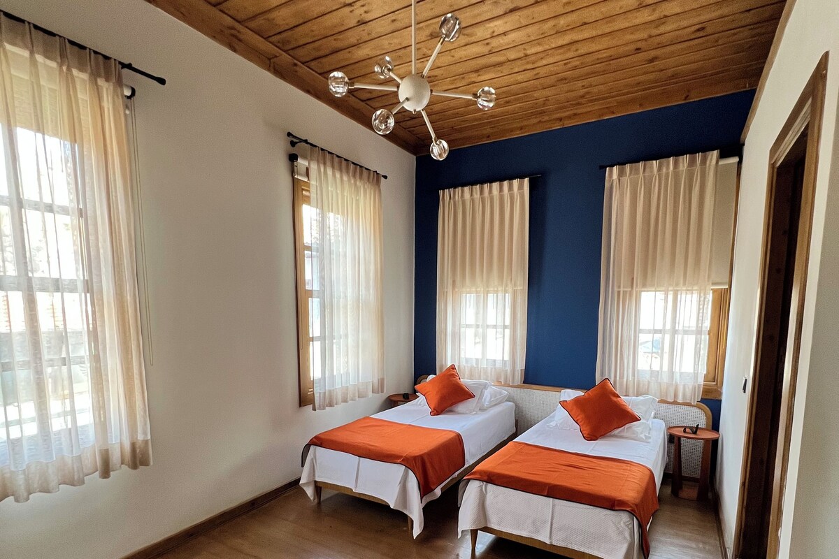 A room with two beds, wooden flooring, a ceiling fan, and large windows with curtains.