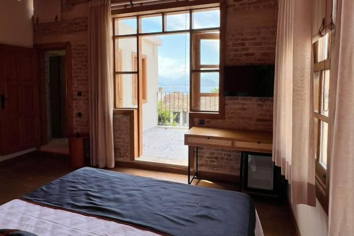 A room with a bed, a desk, and a TV. There is a large window with curtains, and a view of the outside.