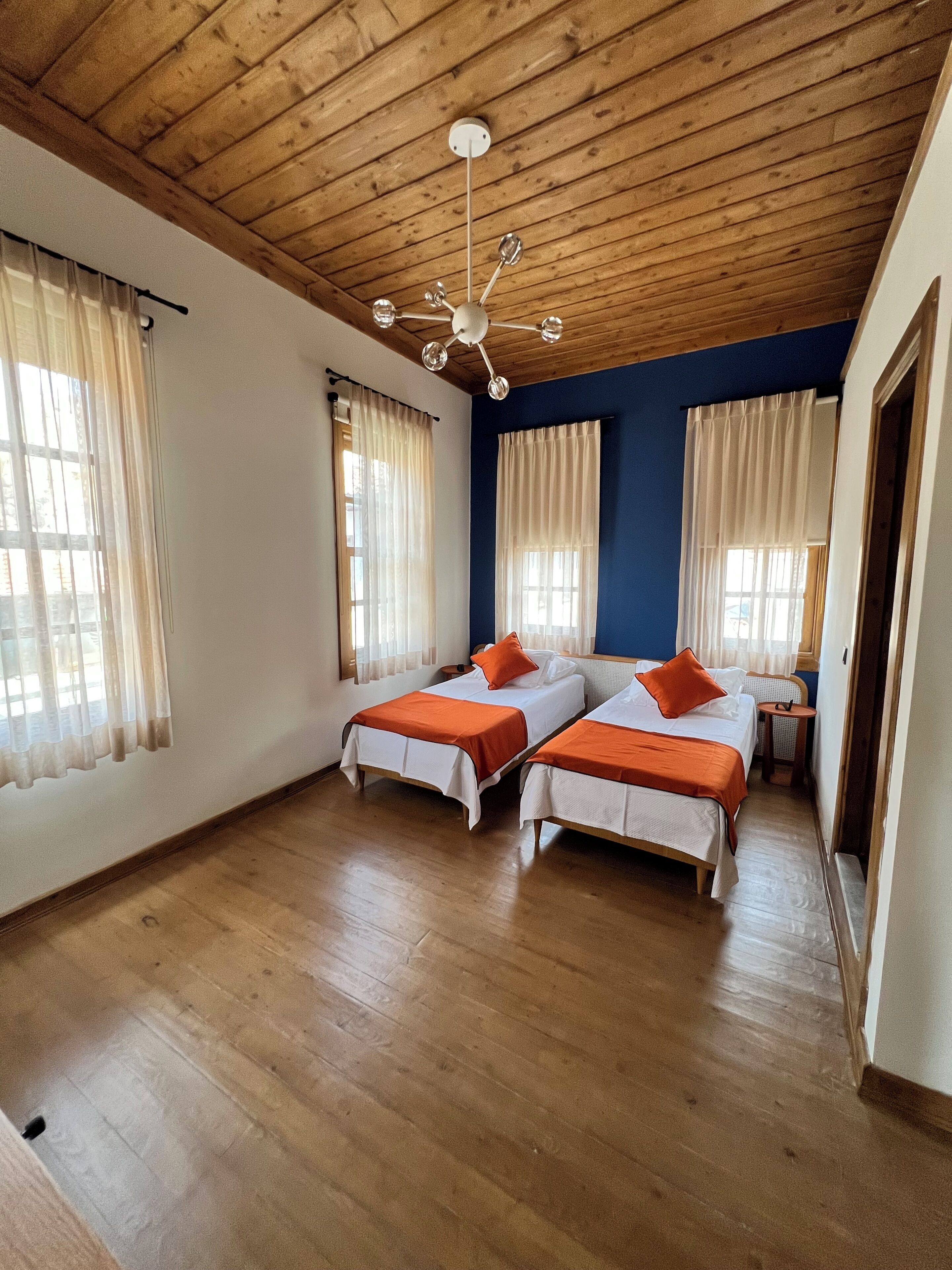 Ein Zimmer mit zwei Betten, Holzboden, Deckenventilator und großen Fenstern mit Vorhängen.