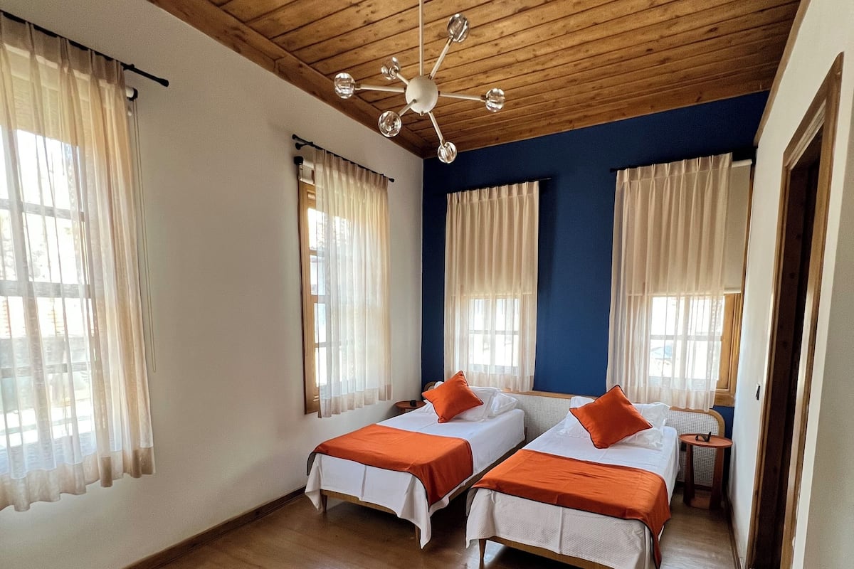 A room with two beds, wooden flooring, a ceiling fan, and large windows with curtains.