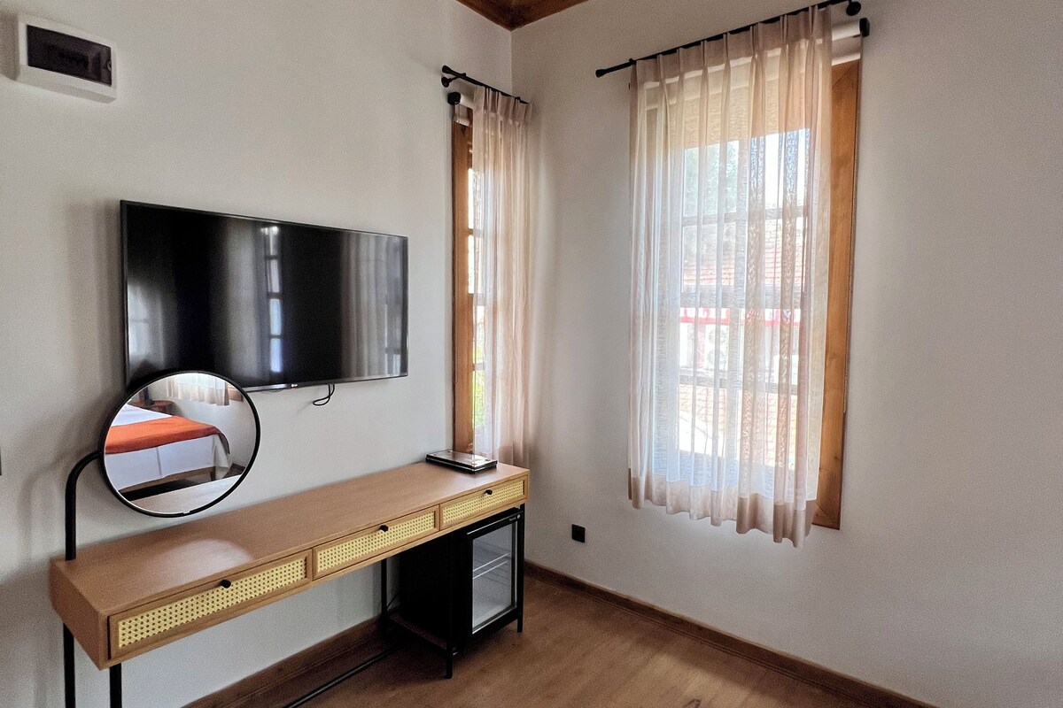 A room with a wooden ceiling, a flat-screen TV mounted on the wall, a desk with drawers, a small cabinet, and a window with sheer curtains.