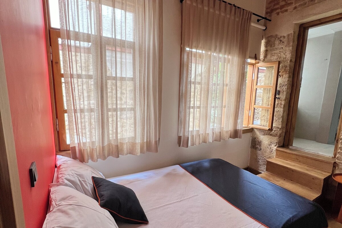 A bedroom with a bed, a window with curtains, a wooden bench, and a stone wall.